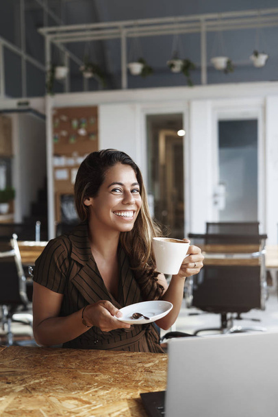 Vertical shot gorgeous hispanic woman taking coffee break, sit alone co-working space or cafe, laptop opened on table, she holds cup as tastes delicious cappuccino, smiling camera happy - Foto, Bild