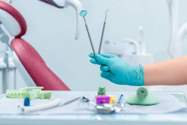 Female hand of dentist in blue gloves holding mouth mirror and periodontal explorer scaler, blurred background of clinic with dental unit, dentistry and teeth care concept with copy space - Foto, Bild