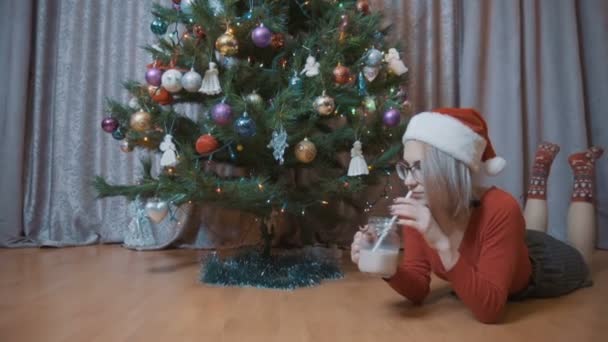 The girl in red is lying on his stomach and drinking cocoa at the Christmas tree. Girl in christmas hat - Video, Çekim