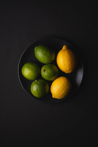 Lime and lemon sour fruits in black plate on moody dark background, top view, vitamins and healthy food - Фото, зображення