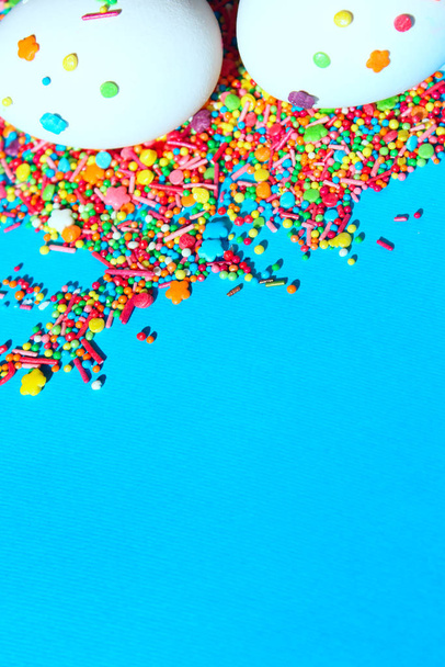 Vertikální oříznutý obraz bílých vajec a roztroušených barevných bonbónů na modrém pozadí. Velikonoce, jídlo. Barevné jídlo pozadí. - Fotografie, Obrázek