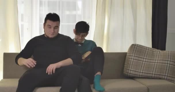 Annoying roommates coming to focused Caucasian boy reading and sitting down on couch on both sides. University students bullying nerd guy in eyeglasses. Cinema 4k ProRes HQ. - Imágenes, Vídeo