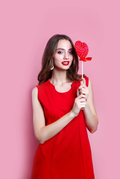 Beautiful brunette girl in red dress holding a red heart-shaped lollipop on pink background. Valentines Day - Фото, зображення