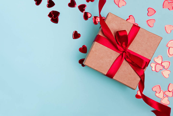 Giftbox with red ribbon and heart shaped confetti on blue background. - Foto, afbeelding
