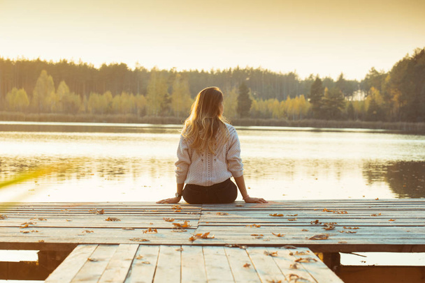 A young slender blonde sits on a wooden pier with her back . - Foto, Bild