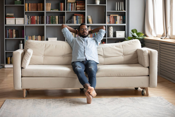 Serene barefoot african man resting on sofa hands behind head - Foto, imagen