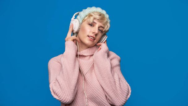 Happy young woman in headphones listening to music, wearing winter knitted hat and sweater on blue background - 写真・画像