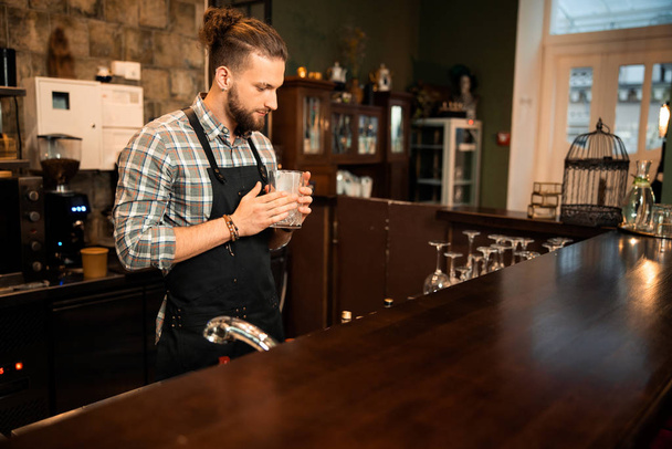 Красивый бородатый бармен в фартуке держит стакан
 - Фото, изображение