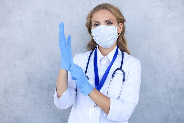 Young woman doctor with stethoscope putting a medical gloves, isolated on grey background. - Foto, immagini