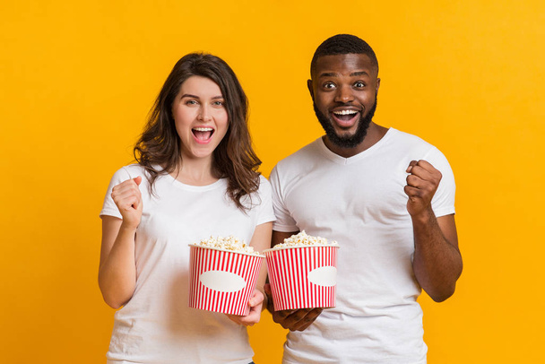 Joyful interracial couple cheering with popcorn buckets, raising fists in excitement - 写真・画像