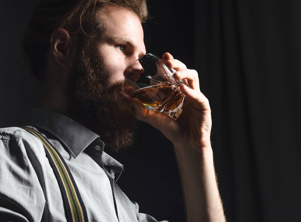 Handsome young man with a beard drinks alcohol from a glass on an isolated black background - Foto, Imagen