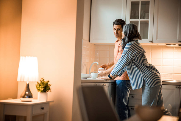 Smiling man looking at woman while washing dishes - Foto, imagen