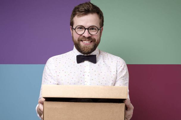 Homme drôle dans des lunettes rondes tient une boîte avec un cadeau dans les mains, il sourit et regarde joyeusement la caméra
. - Photo, image