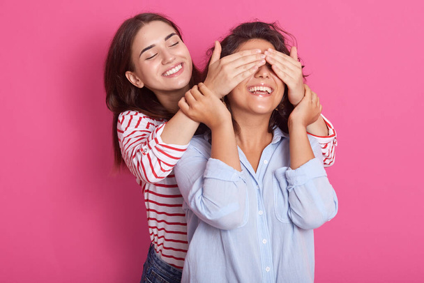 Close up portrait of beautiful Caucasian woman covering eyes to her friend standing behind and smiling, looking at girl wears blue casual shirt, posing isolated on pink background. Friendship concept. - Fotoğraf, Görsel