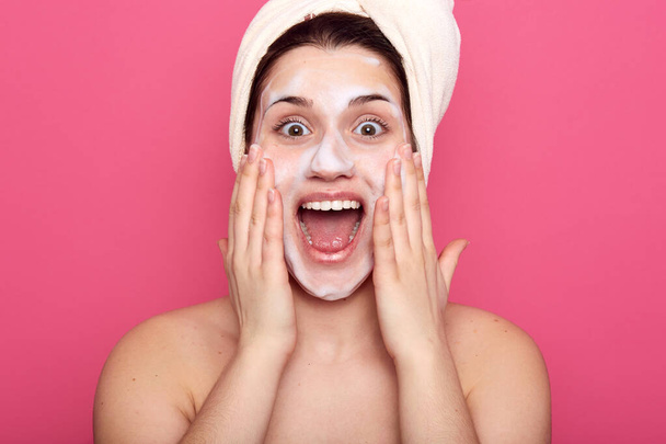 Studio shot of pretty girl with facial mask, surprised female with white towel on her head, lady having astonished expression, posing isolated over pink background. Skin care and beauty concept. - Zdjęcie, obraz