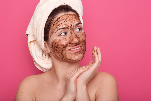 Close up portrait of girl wrapped hair with towel, posing with skin covered by mask. Model with brown face, doing morning procedures, looking dreamily aside, standing with bare shouldesat home. - Foto, imagen