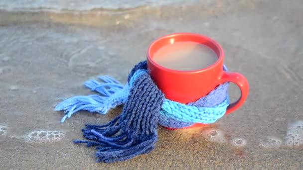 Caneca vermelha com café amarrado com cachecol de malha azul fica na praia de areia
 - Filmagem, Vídeo