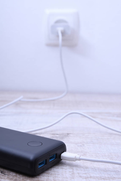 Black phone power bank plugged in the socket on the wall for charging on wooden background - Photo, Image