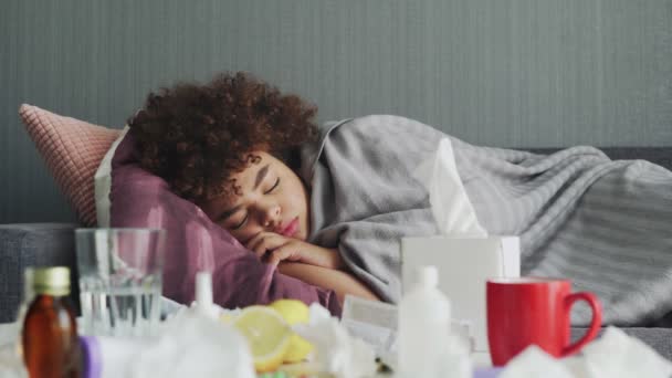 Ill νεαρή Αφροαμερικανή κοπέλα κοιμάται χαλαρωτικό κατά τη διάρκεια της ασθένειας που βρίσκεται στον καναπέ στο διαμέρισμα κάτω από ζεστό καρό. - Πλάνα, βίντεο