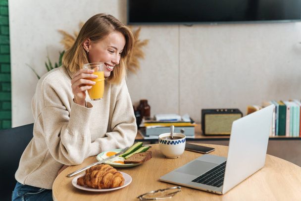 Photo of joyful blonde woman using laptop while having breakfast - Foto, afbeelding