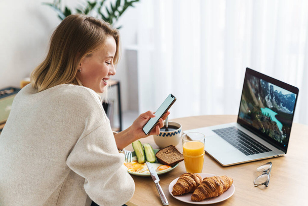 Photo of smiling woman using laptop and cellphone while having breakfast - Foto, Bild