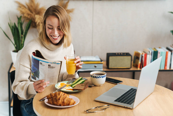 Photo of smiling blonde woman reading magazine while having breakfasta - Foto, afbeelding