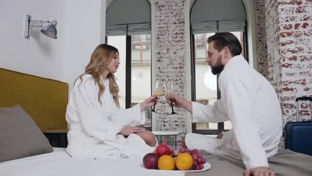 Adorable happy amorous young couple in bathrobes sitting on bed and drinking champagne in the beautifully designeted hotel room - Materiał filmowy, wideo