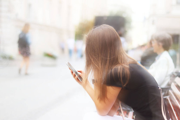 Молода красива дівчина дивиться в смартфон, який тримає. Жінка сидить на лавці в центрі стародавнього міста перехожих в розмитому розмитому. Стиль життя та диверсії сучасної молоді
 - Фото, зображення
