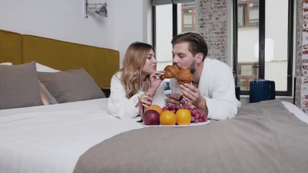 Front view of attractive happy smiling young couple in love dressed in bathrobes which lying on bed and girl feeding her boyfreind with croissant - Кадры, видео