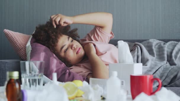 Nemocná africká americká dívka s chřipkou ležící doma na gauči. Nemocná mladá černoška se studenou kontrolní teplotou s teploměrem - Záběry, video
