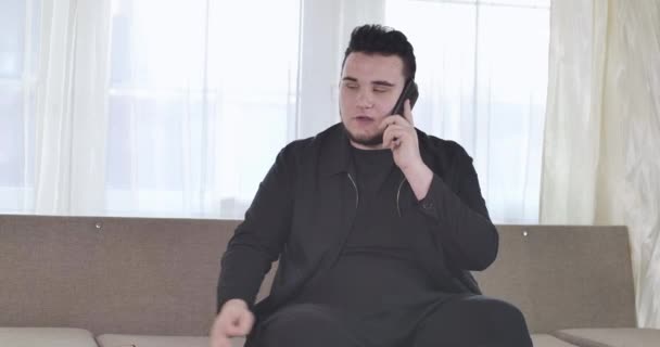 Portrait of confident Caucasian plump man talking on the phone emotionally. Serious brunette guy sitting on couch indoors and using smartphone. Lifestyle, communication. Cinema 4k ProRes HQ. - Záběry, video
