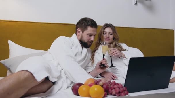 Close view of happy modern amorous young couple in white bathrobes which lying on bed and drinking sparkling wine in hotel room - Materiaali, video