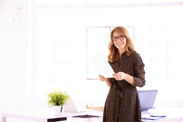 Tiro de atractiva mujer de negocios de mediana edad sonriente usando vestido mientras está de pie en el escritorio de la oficina
.  - Foto, imagen