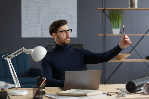 Architect working in office with laptop.Engineer thinks over architectural plan, searching new ideas for construction project. Portrait of handsome bearded man sitting at workplace. Business concept - Foto, imagen