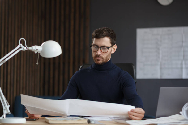 Architect working in office with blueprints.Engineer inspect architectural plan, sketching a construction project.Portrait of handsome bearded man sitting at workplace. Business construction concept - 写真・画像