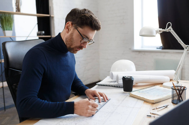 Architect working in office with blueprints.Engineer inspect architectural plan, sketching a construction project.Portrait of handsome bearded man sitting at workplace. Business construction concept - Valokuva, kuva