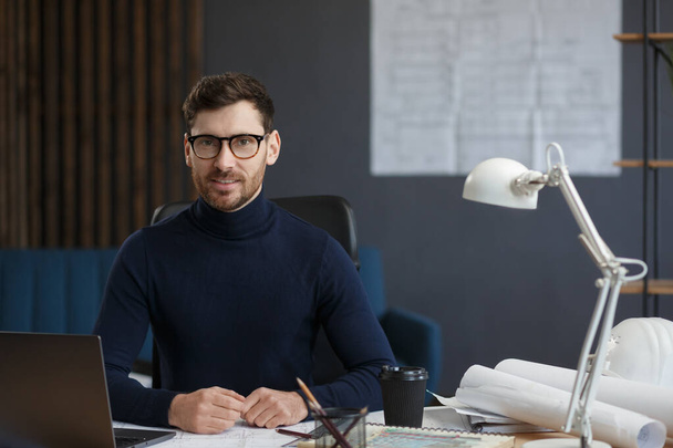 Architect working in office with laptop. Business portrait of handsome bearded man wearing eyeglasses sitting at workplace. Confident businessman became successful. Business concept - Foto, afbeelding