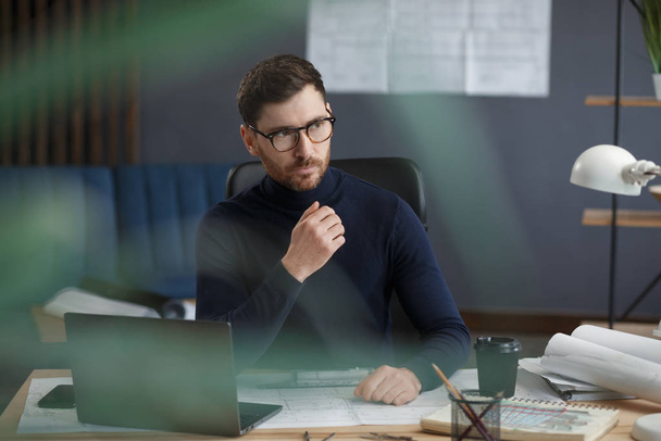 Architect working in office with laptop. Business portrait of handsome bearded man wearing eyeglasses sitting at workplace. Confident businessman became successful. Business concept - Фото, изображение