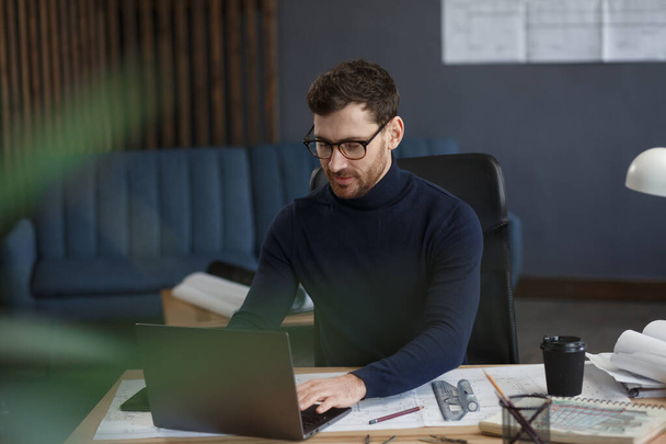 Architect working in office with laptop. Engineer thinks over architectural plan, searching new ideas for construction project. Portrait of handsome bearded man sitting at workplace. Business concept - Photo, image