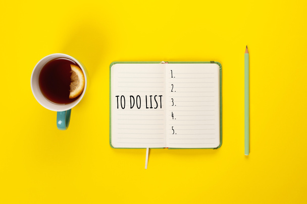 Zrobić listę. Miętowy kubek herbaty i nowoczesny notatnik z listy do zrobienia na jasnożółtym biurku z góry - Zdjęcie, obraz