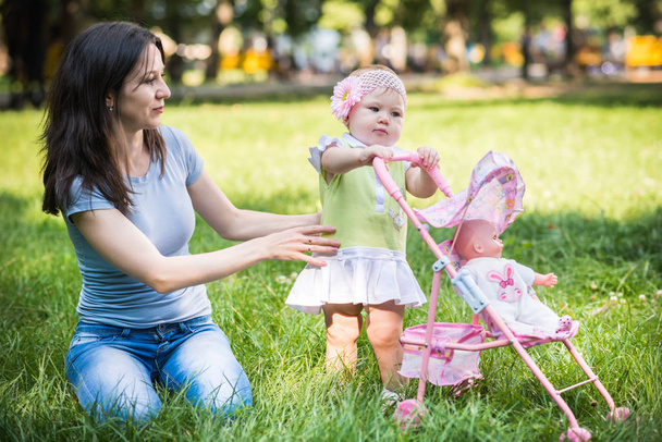 Mother sitting on grass and helping daughter to walk and roll toy stroller - Φωτογραφία, εικόνα