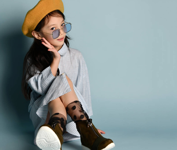 Little asian child in sunglasses, shirt dress, brown beret, boots. Smiling, posing sitting on blue background. Close up - Valokuva, kuva