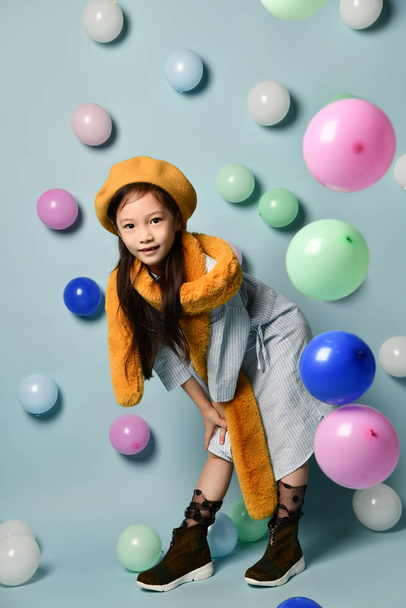 portrait of a little Asian woman in a denim dress and a stylish scarf with a beret., with a wide toothless smile against the background of many multi-colored balloons. - Photo, image
