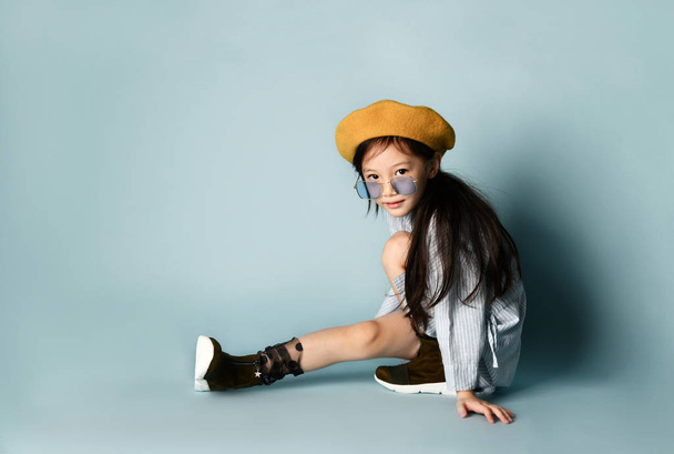 Little asian kid in sunglasses, shirt dress, brown beret, boots. Smiling, posing sitting sideways on blue background. Close up - Foto, afbeelding