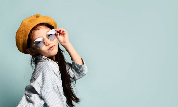 Little asian kid in sunglasses, oversized shirt dress, brown beret, boots. She kissing you, posing on blue background. Full length - Zdjęcie, obraz