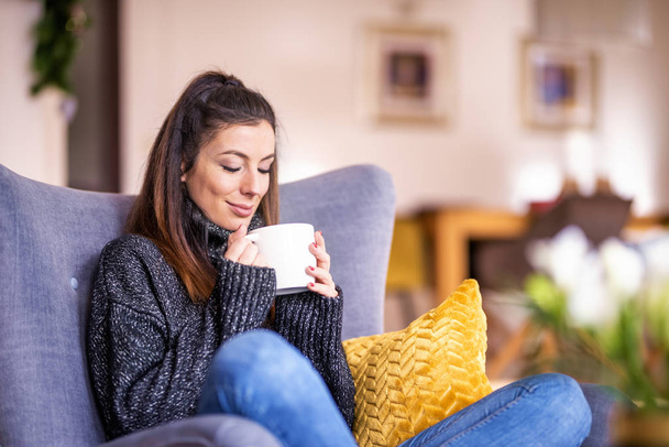 Smiling woman wearing turtleneck sweater and jeans while relaxing in the armchair at home and drinking tea. - 写真・画像