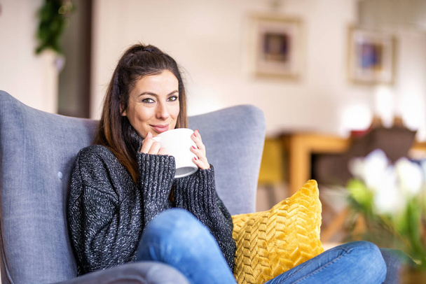 Smiling woman wearing turtleneck sweater and jeans while relaxing in the armchair at home and drinking tea. - Φωτογραφία, εικόνα