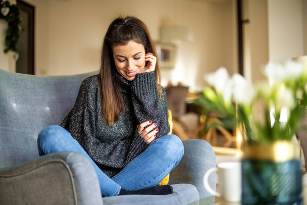 Shot of an attractie smiling woman wearing turtleneck sweater and jeans while relaxing in the armchair at home. - Foto, imagen