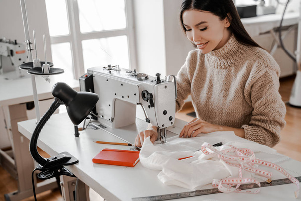 縫製産業では、縫製機械のアジアの女性が服をモデル化しています。縫製、デザイン作業、テーラリングスタジオ、テーラーリング、デザイナー服、製造、創造的な開発の過程で - 写真・画像