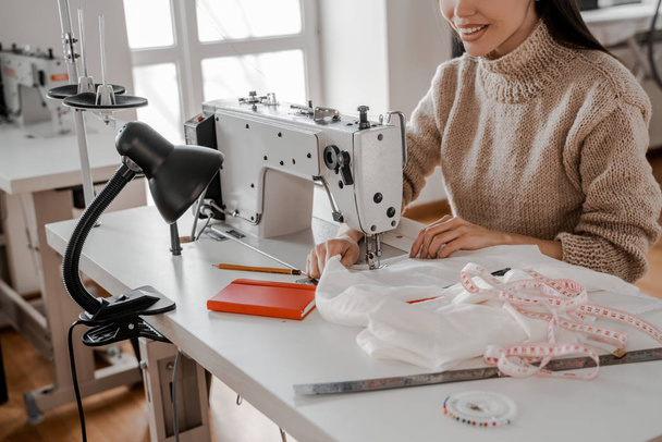 The woman smiles and sews at the industrial level with a sewing machine. Sewing, design work, tailoring studio, tailor, designer clothes, manufactory, in the process of creative development - Fotoğraf, Görsel